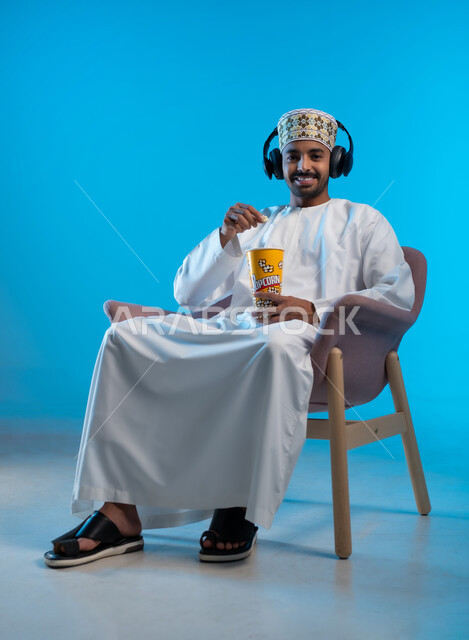 التسلية والترفيه بقضاء أوقات ممتعة ومسلية، الاستمتاع بمشاهدة الأفلام، مواهب غنائية فنية، بورتريه لشاب عربي خليجي عماني يرتدي الدشداشه والكمه يجلس على كرسي مريح يمسك بيده علبة الفوشار، الانصات للأغاني بإستخدام الهيدفون، خلفية زرقاء