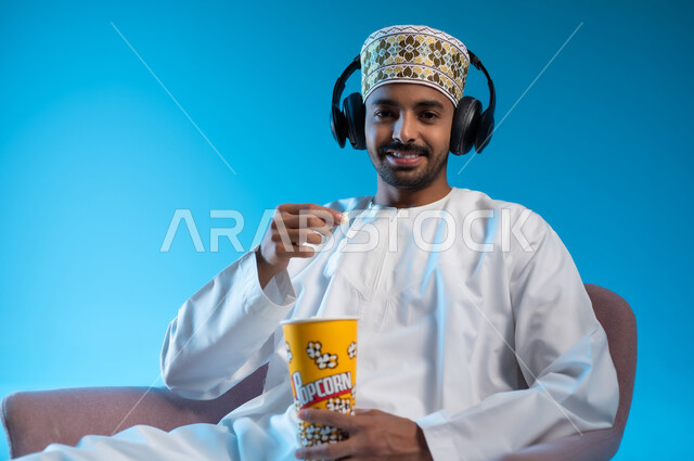 الاستمتاع بمشاهدة الأفلام، التسلية والترفيه بقضاء أوقات ممتعة ومسلية، مواهب غنائية فنية، بورتريه لشاب عربي خليجي عماني يرتدي الدشداشه والكمه يجلس على كرسي مريح يمسك بيده علبة الفوشار، الانصات للأغاني بإستخدام الهيدفون، خلفية زرقاء