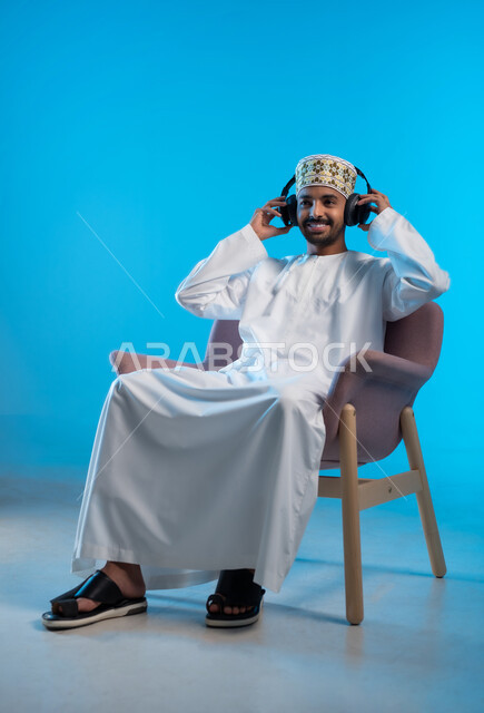 تعابير الاندماج مع الأغنية، مواهب غنائية فنية، بورتريه لشاب عربي خليجي عماني يرتدي الدشداشه والكمه يجلس على كرسي مريح ينظر الى الكاميرا يستمع الى الموسيقى من خلال سماعه الرأس، الانصات والإستماع للأغاني بإستخدام الهيدفون، التفاعل مع الانغام الموسيقية، خلفية زرقاء