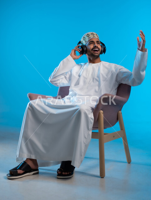 مواهب غنائية فنية، إيماءات وتعابير الاندماج مع الأغنية، بورتريه لشاب عربي خليجي عماني يرتدي الدشداشه والكمه يجلس على كرسي مريح يستمع الى الموسيقى من خلال سماعه الرأس، الانصات والإستماع للأغاني بإستخدام الهيدفون، التفاعل مع الانغام الموسيقية، خلفية زرقاء