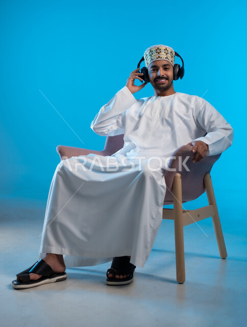 تعابير الاندماج مع الأغنية، مواهب غنائية فنية، بورتريه لشاب عربي خليجي عماني يرتدي الدشداشه والكمه يجلس على كرسي مريح ينظر الى الكاميرا يستمع الى الموسيقى من خلال سماعه الرأس، الانصات والإستماع للأغاني بإستخدام الهيدفون، التفاعل مع الانغام الموسيقية، خلفية زرقاء