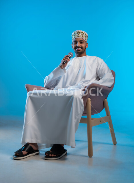 تعابير الاندماج والتفاعل مع الموسيقا، تسجيل مقطع صوتي غنائي، بورتريه لشاب عربي خليجي عماني مبتسم يرتدي الكمة و الدشداشة يمسك بيده المايكروفون يقوم بالغناء ينظر الى الكاميرا بإيماءات الفرح والسرور، تقديم العروض الصوتية، خلفية زرقاء