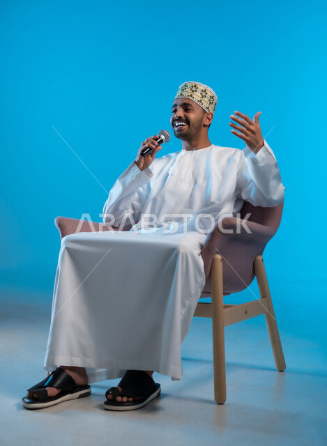 تسجيل مقطع صوتي غنائي، تعابير الاندماج والتفاعل مع الموسيقا، بورتريه لشاب عربي خليجي عماني مبتسم يرتدي الكمة و الدشداشة يمسك بيده المايكروفون يقوم بالغناء بإيماءات الفرح والسرور، تقديم العروض الصوتية، خلفية زرقاء