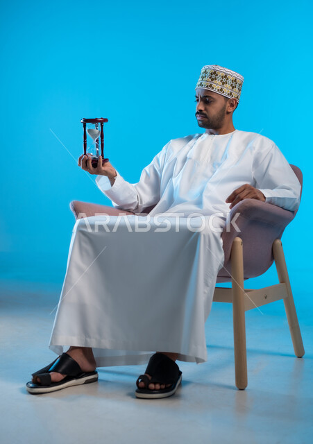 مفهوم إدارة الأعمال وتنظيم الجدول الزمني, القيام بمراقبة توقيت الرمل, إدراك قيمة اللحظات الثمينة, بورتريه لشاب عربي خليجي عماني يرتدث الكمة و الدشداشة يجلس على كرسي مريح يحمل بيده الساعة الرملية الزجاجية (البنكام) وينظر إليها, خلفية زرقاء