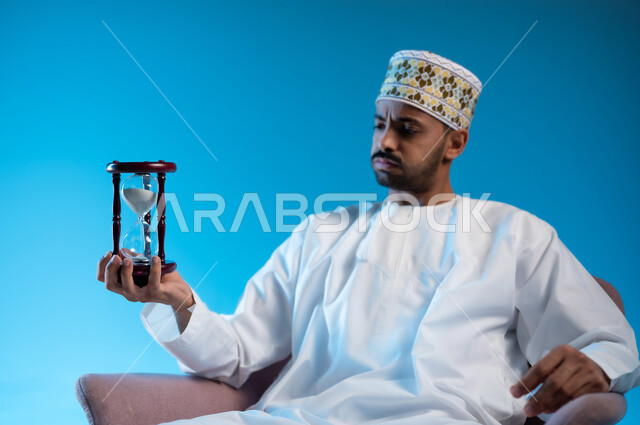 إدراك قيمة اللحظات الثمينة, القيام بمراقبة توقيت الرمل, مفهوم إدارة الأعمال وتنظيم الجدول الزمني, بورتريه لشاب عربي خليجي عماني يرتدي الكمة و الدشداشة يجلس على كرسي مريح يمسك بيده الساعة الرملية الزجاجية (البنكام) وينظر إليها, خلفية زرقاء