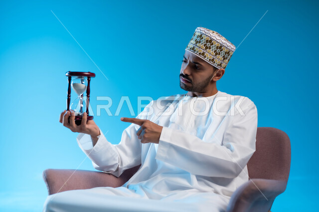 القيام بمراقبة توقيت الرمل, إدراك قيمة اللحظات الثمينة, مفهوم إدارة الأعمال وتنظيم الجدول الزمني, بورتريه لشاب عربي خليجي عماني يرتدي الكمة و الدشداشة يجلس على كرسي مريح يشير بأصبع السبابة الى الساعة الرملية الزجاجية (البنكام) وينظر إليها, خلفية زرقاء
