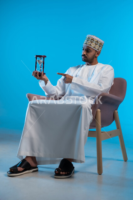 القيام بمراقبة توقيت الرمل, إدراك قيمة اللحظات الثمينة, مفهوم إدارة الأعمال وتنظيم الجدول الزمني, بورتريه لشاب عربي خليجي عماني يرتدي الكمة و الدشداشة يجلس على كرسي مريح يشير بأصبع السبابة الى الساعة الرملية الزجاجية (البنكام) وينظر إليها, خلفية زرقاء