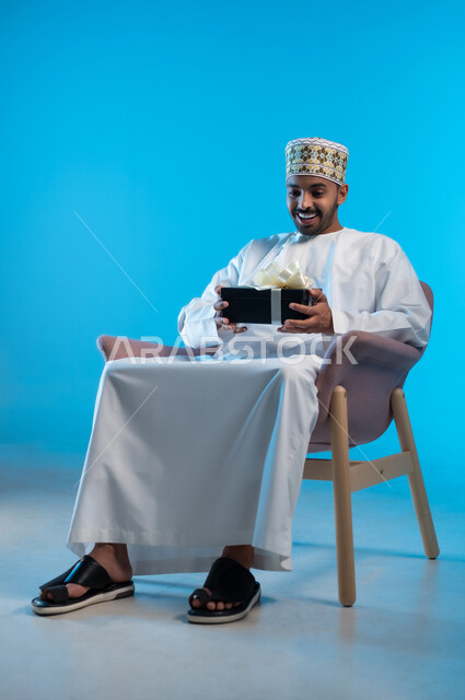 تقديم الهدايا بالاعياد والمناسبات الخاصة، مفهوم الشكر والامتنان، التعبير عن الحب والتقدير والاحترام، بورتريه لشاب عربي خليجي عماني يرتدي الكمة و الدشداشة يجلس على كرسي مريح يحمل بيديه صندوق هدية, ايماءات الفرح والسرور، خلفية زرقاء