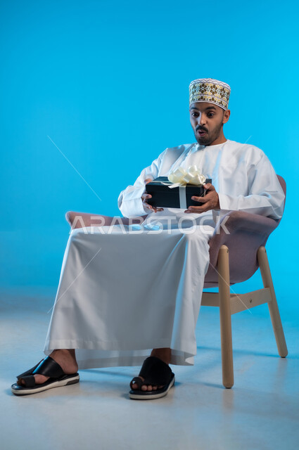 The concept of thanks and gratitude, giving gifts on holidays and special occasions, expressing love, appreciation and respect, a portrait of a young Gulf Arab Omani man wearing a turban and a dishdasha sitting on a comfortable chair holding a gift box in his hands, gestures of surprise and shock, blue background