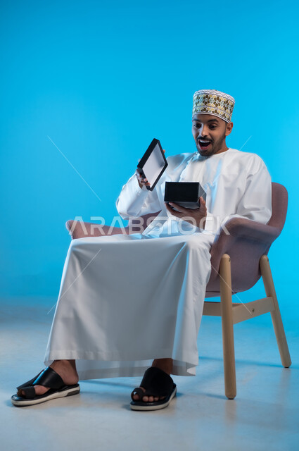 مفهوم الشكر والامتنان، تقديم الهدايا بالاعياد والمناسبات الخاصة، التعبير عن الحب والتقدير والاحترام، بورتريه لشاب عربي خليجي عماني يرتدي الكمة و الدشداشة يجلس على كرسي مريح يحمل بيديه صندوق هدية, ايماءات المفاجئة والصدمة، خلفية زرقاء