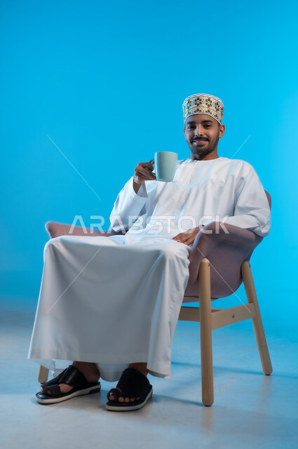 النظر الى الكاميرا بإيماءات السعادة و الفرح, قضاء وقت الفراغ باحتساء المشروب المفضل, الجلوس والاستمتاع بالمشروبات الساخنة والأعشاب الشتوية الدافئة, بورتريه لشاب عربي خليجي عماني مبتسم يرتدي الكمة و الدشداشة يجلس على كرسي مريح يمسك كوب القهوة, خلفية زرقاء