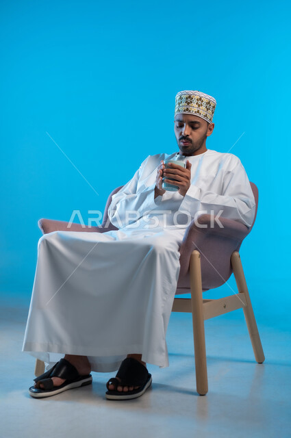 الجلوس والاستمتاع بالمشروبات الساخنة والأعشاب الشتوية الدافئة, قضاء وقت الفراغ باحتساء المشروب المفضل, بورتريه لشاب عربي خليجي عماني مبتسم يرتدي الكمة و الدشداشة يجلس على كرسي مريح يقوم بشرب القهوة بإيماءات السعادة و الفرح, خلفية زرقاء