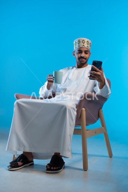 استخدام التقنيات الحديثة المتطورة, الاستمتاع بالمشروبات الساخنة, تصفح مواقع التواصل الاجتماعي عبر الهاتف المحمول, بورتريه لشاب عربي خليجي عماني يرتدي الدشداشة والكمه يجلس على كرسي مريح يحمل بيده كوب من القهوة، خلفيه زرقاء