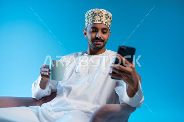 الاستمتاع بالمشروبات الساخنة, تصفح مواقع التواصل الاجتماعي عبر الهاتف المحمول, استخدام التقنيات الحديثة المتطورة, بورتريه لشاب عربي خليجي عماني يرتدي الدشداشة والكمه يجلس على كرسي مريح يحمل بيده كوب من القهوة، خلفيه زرقاء