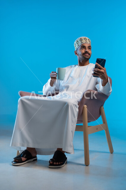 تصفح مواقع التواصل الاجتماعي عبر الهاتف المحمول, الاستمتاع بالمشروبات الساخنة, استخدام التقنيات الحديثة المتطورة, بورتريه لشاب عربي خليجي عماني يرتدي الدشداشة والكمه يجلس على كرسي مريح يحمل بيده كوب من القهوة، خلفيه زرقاء
