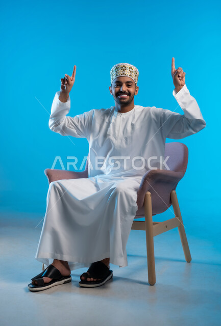 النظر الى الكاميرا بايماءات السعادة والسرور، تحديد موقع معين والإشارة والتوضيح إلى شيء ما، بورتريه لشاب عربي خليجي عمماني يرتدي الكمة والدشداشة يجلس على كرسي مريح يرفع اصبعه السبابة إلى الأعلى، خلفية زرقاء