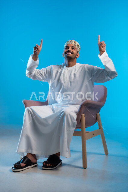 تحديد موقع معين بايماءات السعادة والسرور، الإشارة والتوضيح إلى شيء ما، بورتريه لشاب عربي خليجي عماني يرتدي الكمة والدشداشة يجلس على كرسي مريح يرفع اصبعه السبابة إلى الأعلى، خلفية زرقاء