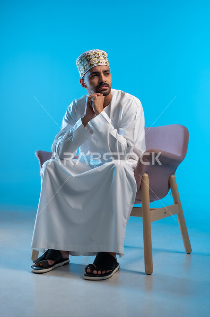 مفهوم شرود الذهن و التأمل والتخطيط للمستقبل, بورتريه لرجل عربي خليجي عماني يرتدي الكمة والدشداشة يجلس على كرسي مريح يضع يده على ذقنه بايماءات التفكير والحيرة، إيجاد حل لمشكلة ما, خلفية زرقاء