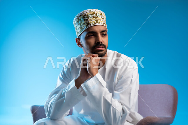 Finding a solution to a problem, the concept of absent-mindedness, contemplation and planning for the future, a portrait of an Arab Gulf Omani man wearing a kimmah and a dishdasha sitting on a comfortable chair, putting his hand on his chin with gestures of thinking and confusion, blue background