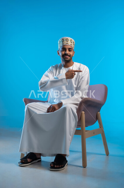 تحديد موقع معين, الإشارة و التوضيح إلى شيء ما, الدلالة على مكان عنصر، بورتريه لشاب عربي خليجي عماني مبتسم يرتدي الكمة والدشداشة يجلس على كرسي مريح ينظر الى الكاميرا يشير بإصبع السبابة إلى شيء ما بإيماءات السرور، خلفية زرقاء