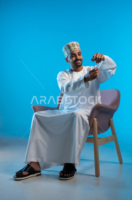 The concept of elegance and masculinity, interest in external appearance, lifting and carrying a certain item, portrait of a young Arab Gulf Omani man wearing a kilt and a dishdasha sitting on a comfortable chair trying to hold something, gestures of happiness and pleasure, blue background