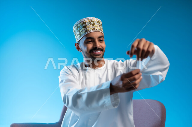 The concept of elegance and masculinity, interest in external appearance, lifting and carrying a certain item, portrait of a young Arab Gulf Omani man wearing a kilt and a dishdasha sitting on a comfortable chair trying to hold something, gestures of happiness and pleasure, blue background
