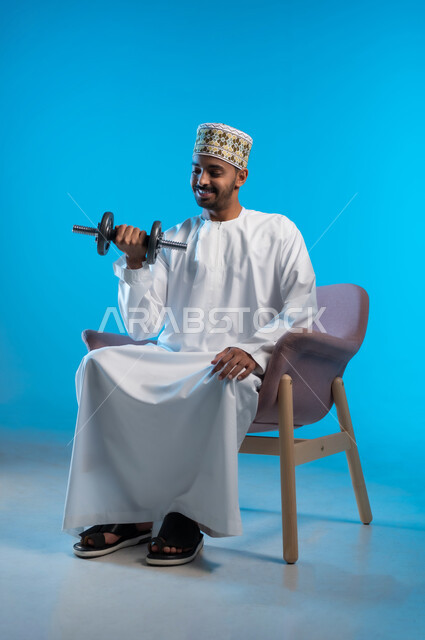 Exercising and lifting weights with dumbbells, portrait of a young Arab Gulf Omani man sitting on a chair wearing a dishdasha and a cloak sitting on a comfortable chair holding heavy iron weights in his hand, maintaining health, physical fitness and physical agility, blue background