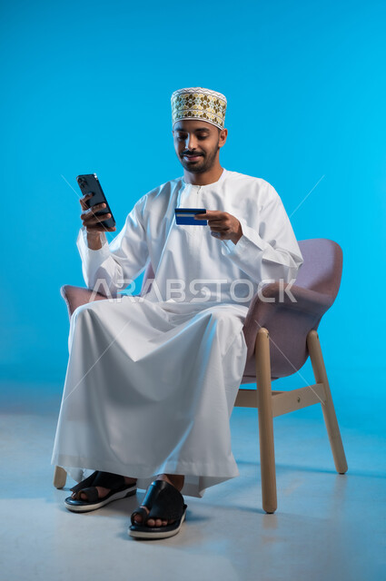 إمكانية التسديد بالبطاقات الإلكترونية, استخدام الأجهزة والتقنيات الحديثة, سهولة الشراء والدفع عن طريق بطاقة الإئتمان البنكية، بورتريه لرجل عربي خليجي عماني يجلس على الكرسي يرتدي الدشداشة و الكمه يمسك بيده الهاتف المحمول, خلفية زرقاء