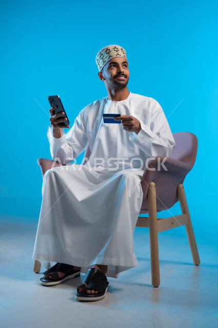 Use of modern devices and technologies, possibility of payment by electronic cards, ease of purchase and payment by bank credit card, an Arab Gulf Omani man sitting on a chair wearing a dishdasha and a sleeve holding a mobile phone in his hand, blue background