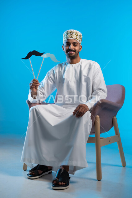 مفهوم الرجولة والقوة، إيماءات البهجة والسرور، بورتريه لشاب عربي خليجي عماني يرتدي الدشداشه والكمه يجلس على كرسي مريح يمسك بيده قصاصة ورقية على شكل شارب، ذكرى اليوم العالمي للرجل ١٩نوفمبر، الاهتمام بالصحة الجسدية للذكور، خلفية زرقاء