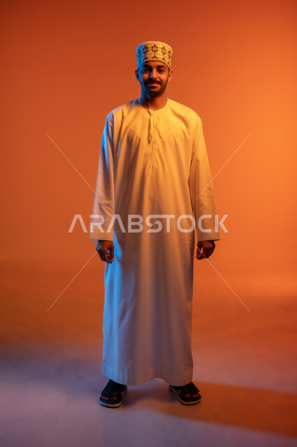 الاهتمام بالأناقة والمظهر الخارجي, مفهوم الرجولة والثقة بالنفس، التقاط الصور الشخصية للوثائق الرسمية، بورتريه لشاب عربي خليجي عماني مبتسم يرتدي الدشداشة و الكمه ينظر الى الكاميرا بإيماءات السرور والسعادة، صورة الجسم بالطول الكامل, خلفية ملونة