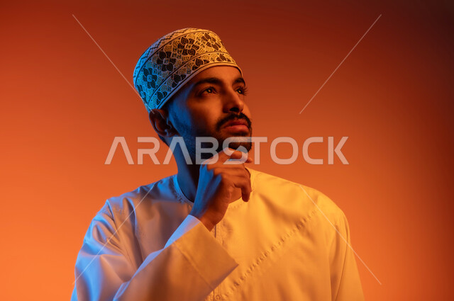مفهوم الرجولة والثقة بالنفس، الاهتمام بالأناقة والمظهر الخارجي, بورتريه لشاب عربي خليجي عماني مبتسم يرتدي الدشداشة و الكمه يضع يده على ذقنه ينظر الى شيء ما بايماءات التفكير والحيرة, خلفية ملونة