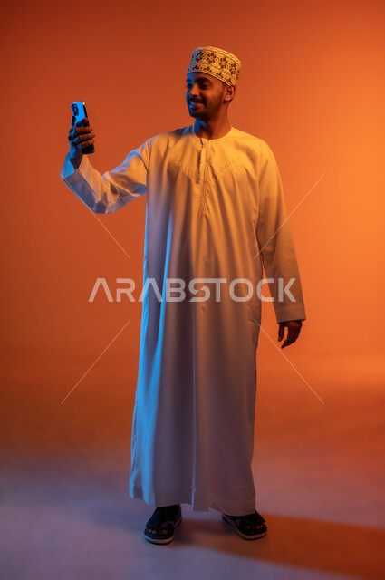 التقاط الصور الشخصية باستخدام الجوال، مفهوم التكنولوجيا الحديثة في تخليد الذكريات، بورتريه لرجل عربي خليجي عماني يرتدي الدشداشة والكمه ويمسك الهاتف المحمول ويأخذ لقطات السيلفي بإيماءات السرور، الجسم بالطول الكامل، خلفية ملونة