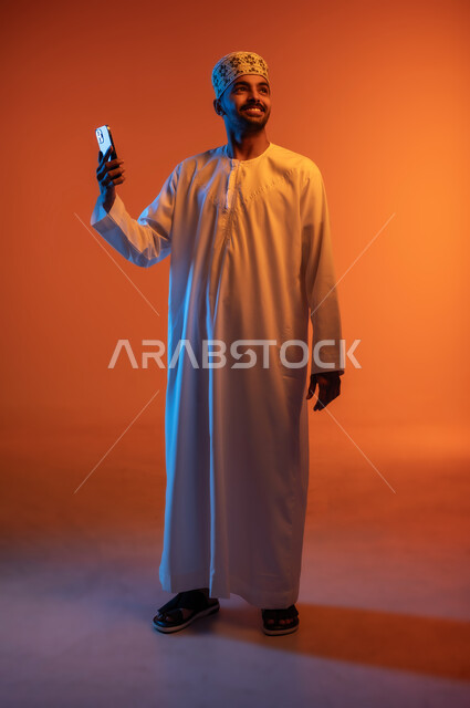 التقاط الصور الشخصية باستخدام الجوال، مفهوم التكنولوجيا الحديثة في تخليد الذكريات، بورتريه لرجل عربي خليجي عماني يرتدي الدشداشة والكمه ويمسك الهاتف المحمول ويأخذ لقطات السيلفي بإيماءات السرور، الجسم بالطول الكامل، خلفية ملونة