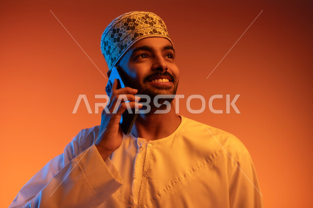 التواصل مع الاهل و الاصدقاء, ايماءات السعادة و السرور، بورتريه لشاب عربي خليجي عماني يرتدي الدشداشه و الكمه يقوم بإجراء مكالمة هاتفية عبر الهاتف المحمول، استخدام التقنيات الحديثة المتطورة, خلفية ملونة
