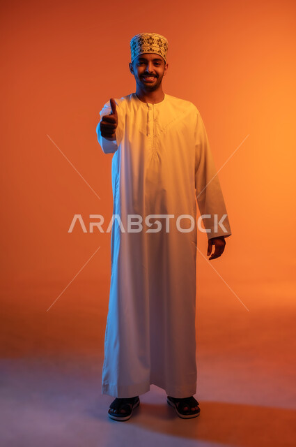 Feeling accepted and satisfied, portrait of a smiling young Arab Gulf Omani man wearing a dishdasha and a sleeve, raising his thumb up with gestures of admiration and pleasure, full body image, colored background