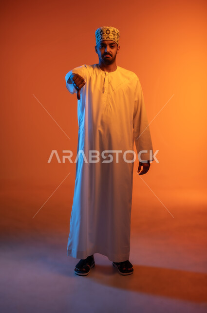 الرفض وعدم القبول، علامات وحركة يد تدل على عدم الإعجاب، النظر إلى الكاميرا بإيماءات الانزعاج والسلبية، بورتريه لشاب عربي خليجي عماني يرتدي الدشداشة والكمه العمانية يشير باصبع الابهام نحو الأسفل، الجسم بالطول الكامل، خلفية ملونة