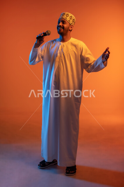 قضاء وقت ممتع بأداء العروض الصوتية، تسجيل مقطع صوتي غنائي, الاستمتاع بممارسة المواهب الفنية، بورتريه لشاب عربي خليجي عماني يرتدي الدشداشة و الكمة يمسك بيده المايكروفون ينظر الى شي ما بايماءات السعادة والسرور, الجسم بالطول الكامل, خلفية ملونة