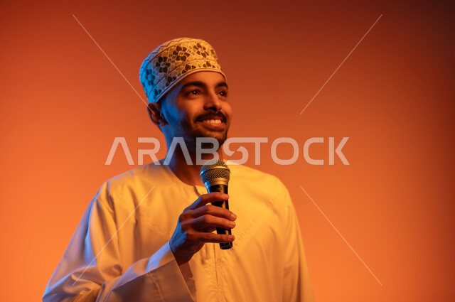 الاستمتاع بممارسة المواهب الفنية، قضاء وقت ممتع بأداء العروض الصوتية، تسجيل مقطع صوتي غنائي,  بورتريه مقرب لشاب عربي خليجي عماني يرتدي الدشداشة و الكمة يمسك بيده المايكروفون ينظر الى شي ما بايماءات السعادة والسرور, خلفية ملونة