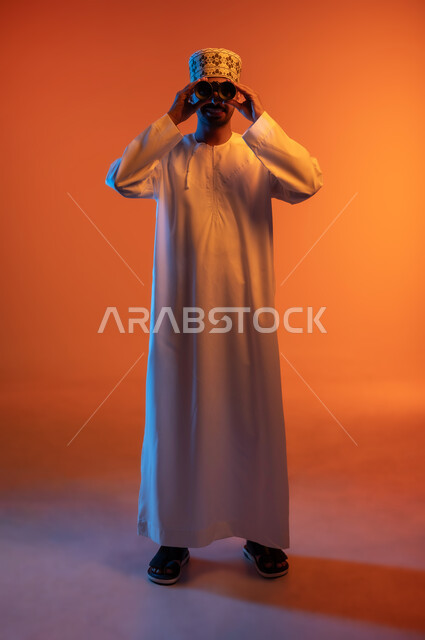 Seeing distant objects through a telescope, love of exploration and adventure, portrait of a smiling young Omani Arab man wearing a dishdasha and a kuma, holding binoculars in his hand and looking at the camera with gestures of enjoyment and pleasure, full-length body image, colored background