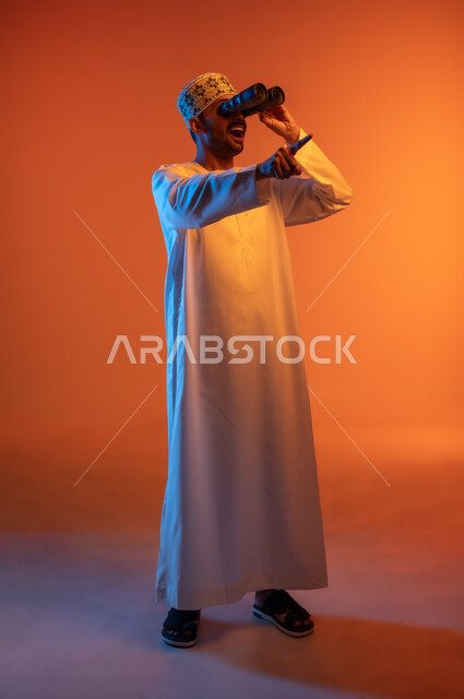 رؤية الاشياء البعيدة عبر التلسكوب, حب الاستكشاف و المغامرة, بورتريه لشاب عربي خليجي عماني مبتسم يرتدي الدشداشة و الكمه يحمل بيده منظار ثنائي العينين يشير باصبع السبابة وينظر الى شيء ما بايماءات الاستمتاع و السرور, صورة الجسم بالطول الكامل, خلفية ملونة