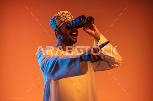 حب الاستكشاف و المغامرة, رؤية الاشياء البعيدة عبر التلسكوب, بورتريه مقرب لشاب عربي خليجي عماني مبتسم يرتدي الدشداشة و الكمه يحمل بيده منظار ثنائي العينين يشير باصبع السبابة وينظر الى شيء ما بايماءات الاستمتاع و السرور, خلفية ملونة