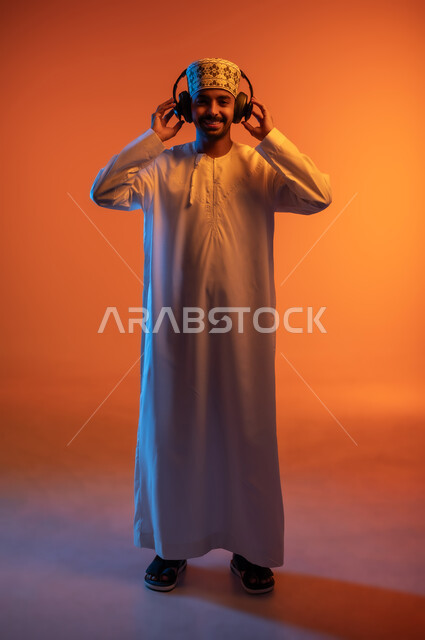 مواهب غنائية فنية، إيماءات الاندماج مع الأغنية، بورتريه لشاب عربي خليجي عماني يرتدي الدشداشه والكمه يستمع الى الموسيقى من خلال سماعه الرأس، الانصات والإستماع للأغاني بإستخدام الهيدفون، التفاعل مع الانغام الموسيقية، صورة الجسم بالطول الكامل، خلفية ملونة