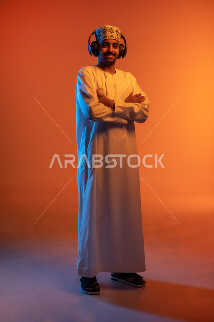 Expressions of integration with the song, artistic singing talents, portrait of a young Arab Gulf Omani man wearing a dishdasha and a kummah with crossed hands listening to music through his headphones, listening and hearing songs using headphones, interacting with musical tunes, full-length body image, colorful background