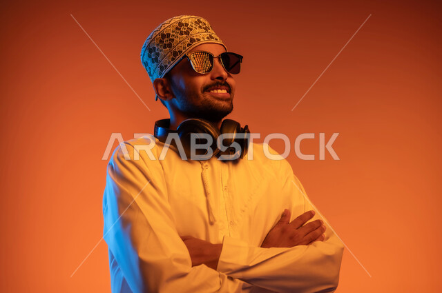 The concept of listening and enjoying, having a good time listening to music with headphones, looking at something with expressions of happiness and pleasure, close-up portrait of a young Arab Gulf Omani man wearing a dishdasha and a kuma standing straight with his hands crossed and wearing his headphones around his neck, colorful background