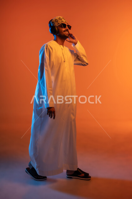 Listening to songs using headphones, expressions of integration with the song, portrait of a young Arab Gulf Omani man wearing a dishdasha, a sleeve and sunglasses looking at something listening to music through his headphones, full-length body image, colored background