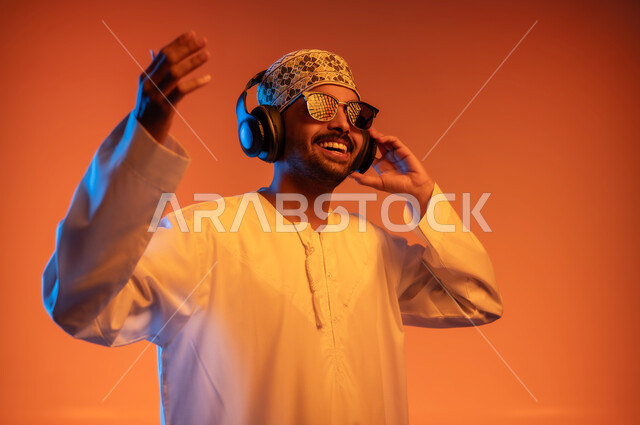 التفاعل مع الانغام الموسيقية، تعابير الاندماج مع الأغنية، بورتريه مقرب لشاب عربي خليجي عماني يرتدي الدشداشه والكمه والنظارة الشمسية يستمع الى الموسيقى من خلال سماعه الرأس، الانصات والإستماع للأغاني بإستخدام الهيدفون، خلفية ملونة