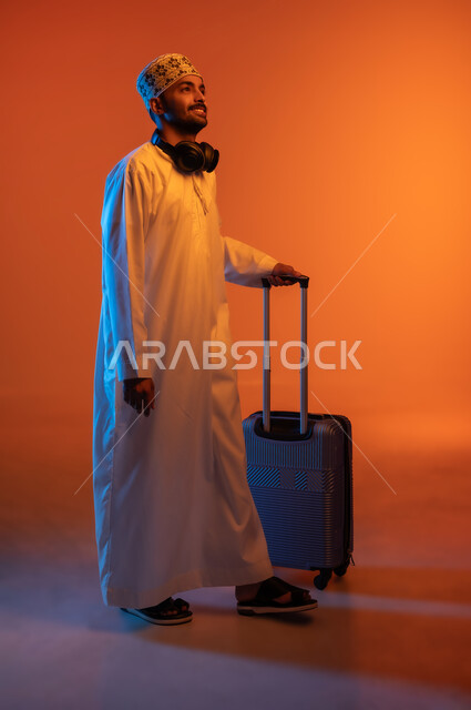 قضاء وقت ممتع بالاستماع الى الموسيقى بواسطة الهيدفون, النظر الى شيء ما بتعابير السعادة والسرور، بورتريه لشاب عربي خليجي عماني يرتدي الدشداشه والكمه يضع سماعه الرأس حول رقبته بجانبه حقيبة سفر، صورة الجسم بالطول الكامل، خلفية ملونة