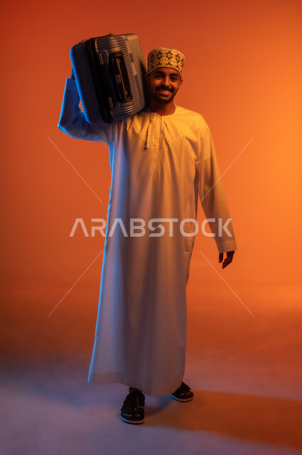 الاستعداد لوقت الرحلة، مفهوم السياحة والسفر، قضاء أوقات ممتعة في الإجازة, بورتريه لشاب عربي خليجي عماني يرتدي الدشداشة و الكمه يحمل حقيبة السفر على كتفه, النظر الى الكاميرا بتعابير الحماس والسرور, خلفيه ملونة