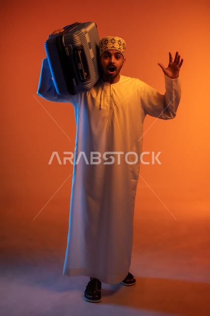 النظر الى الكاميرا بتعابير الصدمة والمفاجئة, الاستعداد لوقت الرحلة، مفهوم السياحة والسفر، قضاء أوقات ممتعة في الإجازة, بورتريه لشاب عربي خليجي عماني يرتدي الدشداشة و الكمه يحمل حقيبة السفر على كتفه, خلفيه ملونة